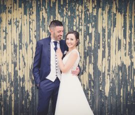 Noivo de fato azul-escuro a olhar para noiva enquanto ela sorri com mão apoiada no peito dele, ambos junto a portão de madeira com tinta descascada em tons de verde e creme, fotógrafo de casamento Santa Maria da Feira Humberto Barbosa